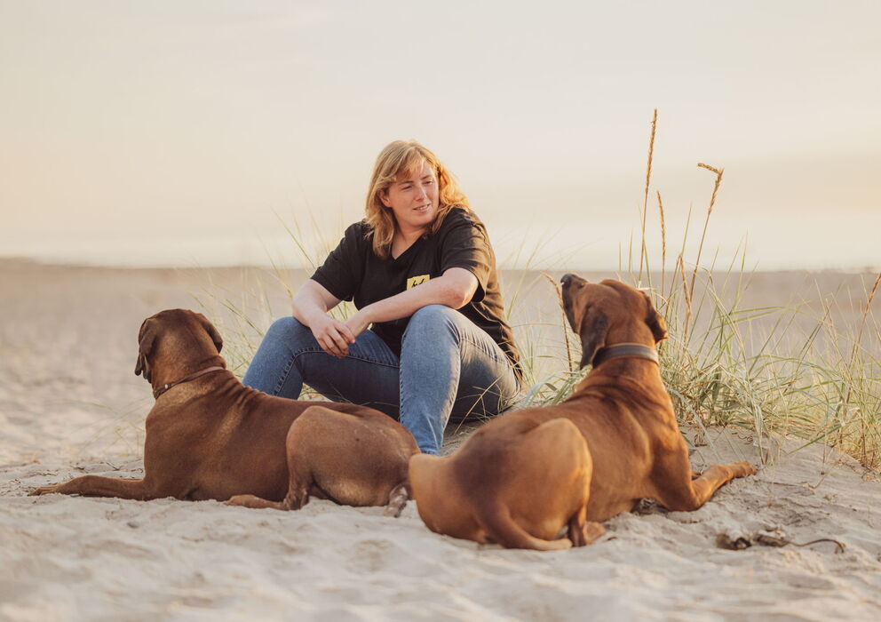 Hundetrainerin Sophia Tumat sitzt am Strand. Vor ihr liegen ihre beiden Ridgeback-Rüden.